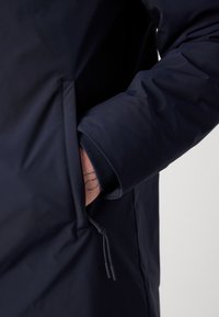 Námořnicky modrá nepromokavá bunda se zipovou kapsou, s hladkou texturou a elastickými manžetami. Tmavá tkanina kontrastuje s detailním prvkem pleti.
