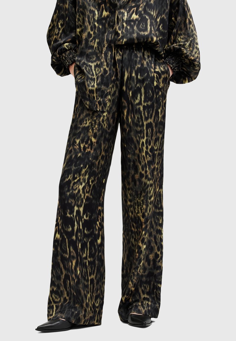 Pantalon ample imprimé léopard et haut assorti portés avec des chaussures noires pointues, mains dans les poches, sur un fond uni.