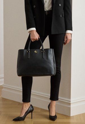 Person i sort blazer og bukser, der holder en stor sort lædertaske med guld RLL-logo, iført sorte høje hæle på trægulv.