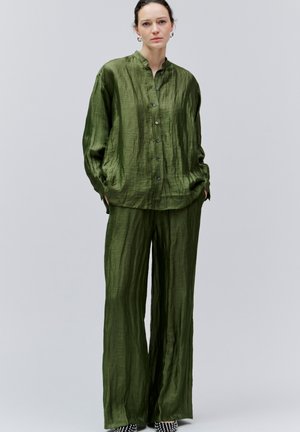 Naine seisab, seljas lahtine oliivirohelise värvusega siidist nööpidega särk ja sobivad laiad püksid, käed taskutes.