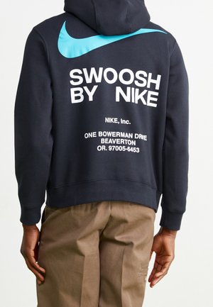 Osoba ubrana w czarną bluzę z kapturem z dużym niebieskim logo Nike i białym napisem "Swoosh by Nike" oraz adresem na plecach.
