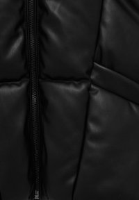 Veste matelassée noire en gros plan, présentant une texture en cuir synthétique, un motif matelassé vertical, et une fermeture éclair centrale avec une finition mate.