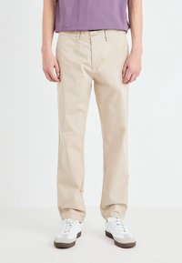 Chinos in cotone beige con design a gamba dritta; dotati di chiusura con bottone, passanti per la cintura e tasche; abbinati a sneakers bianche.