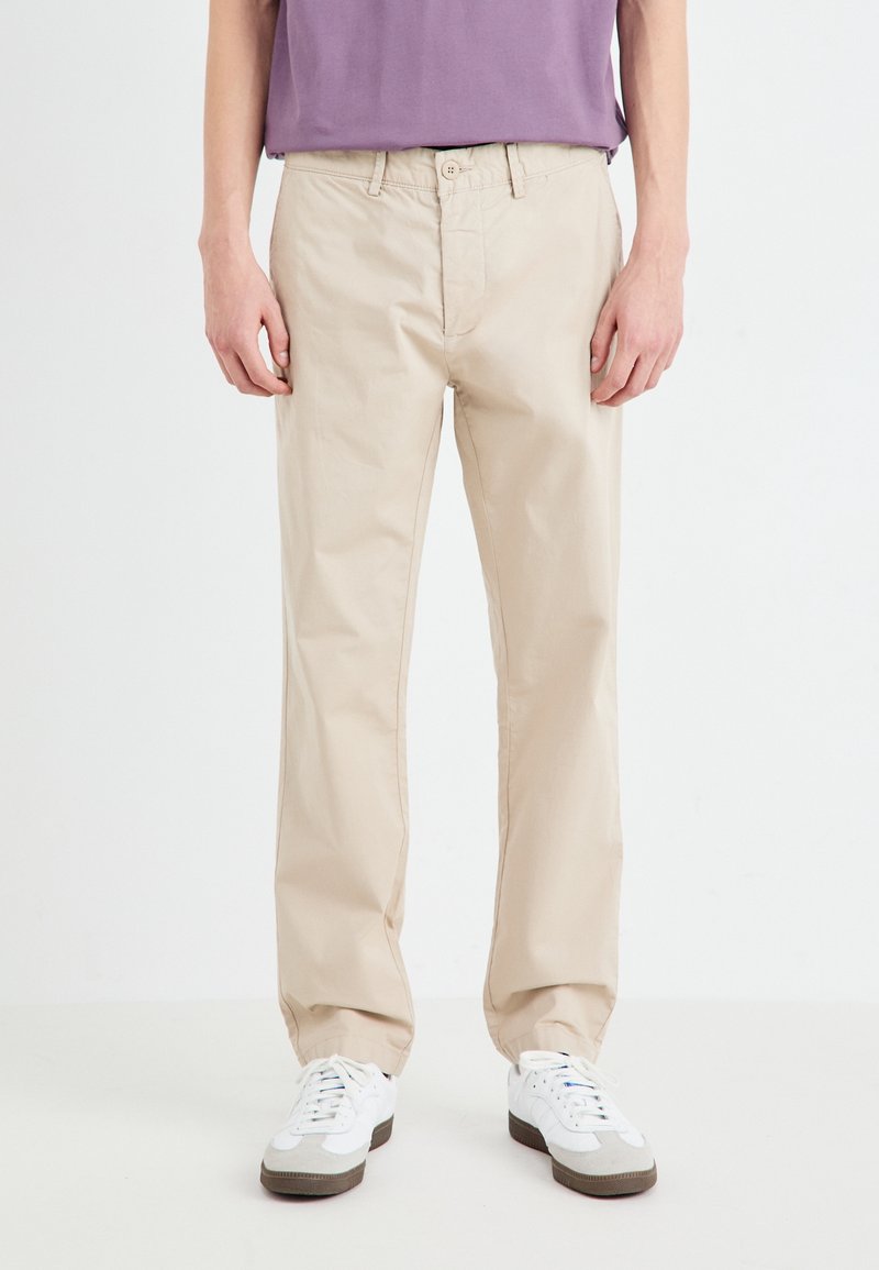 Chinos in cotone beige con design a gamba dritta; dotati di chiusura con bottone, passanti per la cintura e tasche; abbinati a sneakers bianche.
