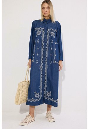 Kvinde iført en lang blå broderet kjole med knapper, der holder en beige vævet taske og har hvide sneakers på.