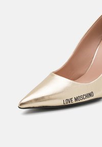 Zlatne metalik visoke potpetice s oštrim vrhom i reljefnim logotipom "LOVE MOSCHINO" sa strane. Glatka tekstura, elegantan dizajn.