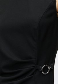Robe noire sans manches avec un anneau en métal argenté à la taille, montrant des fronces et des lignes épurées et modernes.
