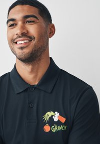 Laivastonsininen poolopaita, jossa on värikäs Grinch-grafiikka vihreine käsineen, punavalkoinen esine ja oranssi pallo. Sileä kangas, klassinen muotoilu.