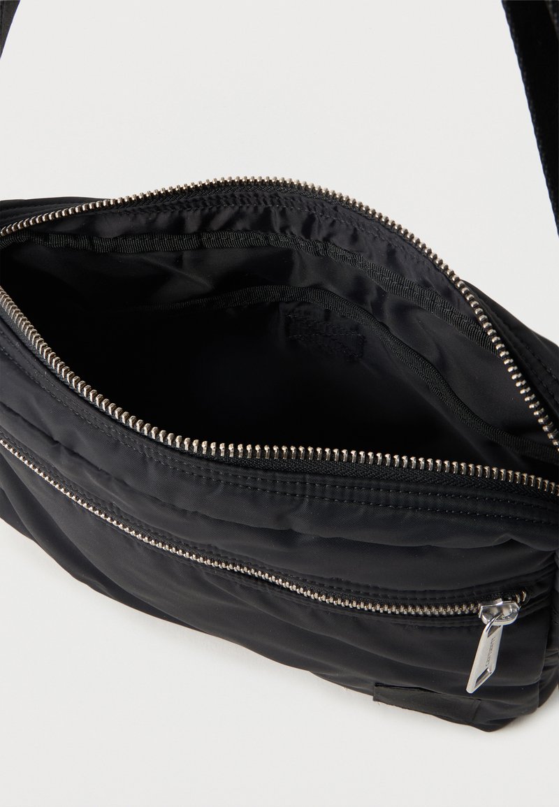 Sac bandoulière en nylon noir avec compartiment principal ouvert et poche zippée à l’avant montrant des détails de fermeture éclair argentés.