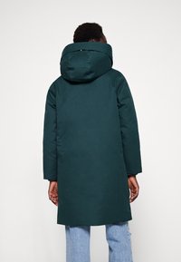 Lang, mørkegrøn hættefrakke med en glat tekstur. Har en afslappet pasform og lange ærmer, båret over lyseblå jeans.