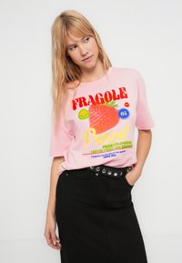 Rožinės spalvos medvilniniai marškinėliai su braškių atvaizdu ir margais užrašais. Trumpomis rankovėmis ir laisvo silueto. Derinami su juoda suknele ir diržu.