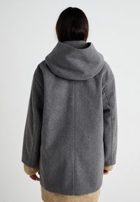 Grå uldfrakke med en rummelig hætte, store ærmer og en strikket beige kant nederst. Minimalistisk design, neutralt farvepalet.