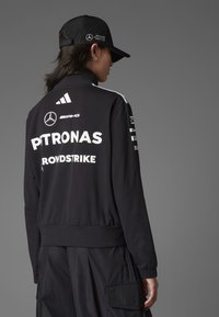 Schwarze Sportjacke mit weißen Logos, hohem Kragen und gerippten Bündchen. Besitzt ein elegantes Design mit seitlichen Streifen. Kombiniert mit einer passenden Kappe.