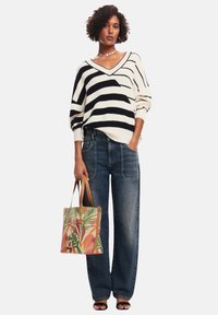 Gestreifter, übergroßer Pullover in Schwarz und Weiß, blaue weit geschnittene Jeans und eine gemusterte Tragetasche mit tropischen Designs, kombiniert mit schwarzen Sandalen.