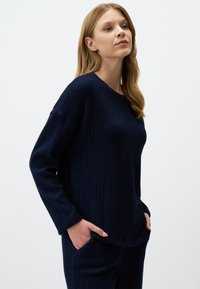 Marineblauer, gestrickter Pullover mit langen Ärmeln, strukturiertem Stoff, Rundhalsausschnitt, seitlichen Taschen und lockerer Passform, seitlich gezeigt.