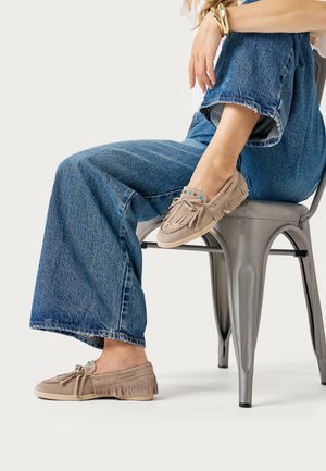 Persoon zittend op een metalen stoel, draagt jeans met wijde pijpen en beige suède mocassins met franjes en turkooizen kralen.