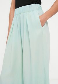 Pantalon à jambes larges bleu clair en tissu doux, doté d'une taille froncée, de plis et de poches latérales. Texture lisse, sans motif.