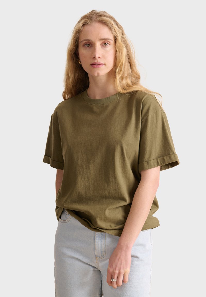 T-shirt in cotone verde oliva con maniche corte e polsini arrotolati, caratterizzato da un colletto rotondo e una vestibilità rilassata. Indossato con jeans azzurri chiari.