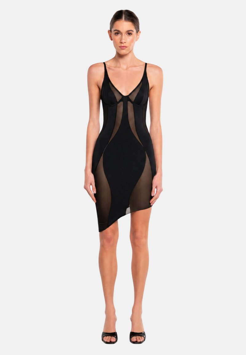 Femme vêtue d'une robe noire ajustée avec des panneaux en maille transparente et des bretelles fines, debout devant un fond clair uni.