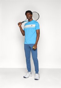 Heledsinine sportlik t-särk, millel on valge horisontaalne triip ja logo, koos keskmise sinise dressipüksiga ja valgete spordijalatsitega, hoiab tennisereketit.