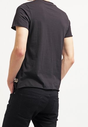 Homme portant un t-shirt noir uni à manches courtes et un pantalon noir, vu de dos avec une main dans la poche.
