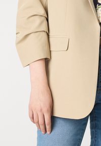 Beiger Blazer mit hochgerollten Ärmeln, der einen sauberen Schnitt und eine glatte Textur aufweist, kombiniert mit hellblauen Jeans.