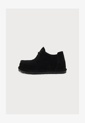 Chaussure décontractée noire en suède avec lacets, semelle épaisse et coutures visibles, montrée de profil sur un fond clair uni.