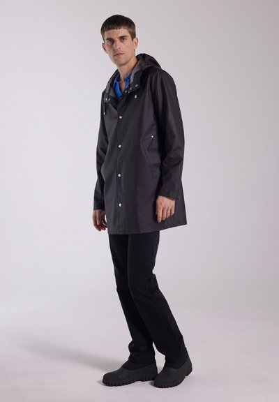 Black waterproof jacket with a hood, silver snap buttons, side pocket, paired with dark jeans and black rubber shoes. Smooth texture.