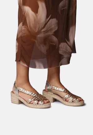 Noge v zlatih tkanih sandalih z odprtimi prsti in blok peto, kombinirane z rjavo cvetlično prosojno midi krilo.