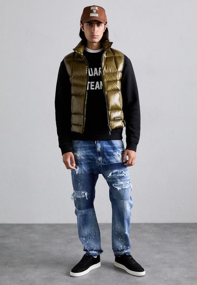 Gilet rembourré vert olive sur un sweat noir avec texte blanc, jeans bleu déchirés et baskets noires. Casquette marron avec texte blanc.