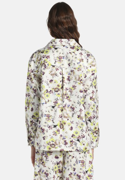 Femme aux longs cheveux bruns portant une chemise blanche à fleurs à manches longues et un pantalon assorti, vue de dos sur un fond uni.