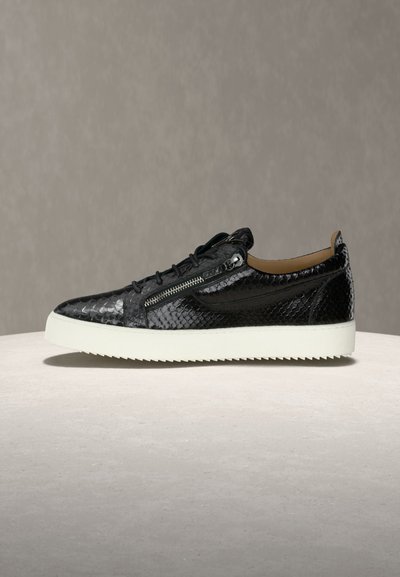 Μαύρο αθλητικό παπούτσι από υφή δέρματος με πλευρικό φερμουάρ, κορδόνια και παχιά λευκή λαστιχένια σόλα σε ουδέτερη γκρι επιφάνεια.