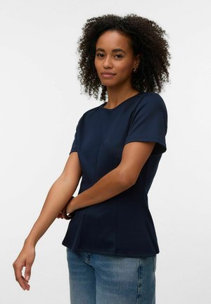 Kvinde med krøllet hår iført en tætsiddende marineblå kortærmet top og blå jeans, der justerer sin venstre ærme mod en ensartet hvid baggrund.