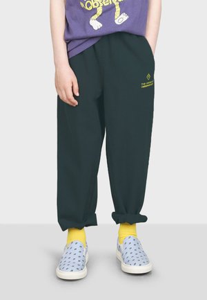 Dunkelgrüne, locker sitzende Jogginghosen mit umgeschlagenen Bündchen und gelbem Logo. Kombiniert mit hellblauen Slippern und gelben Socken.
