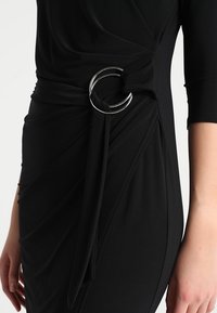 Černé šaty z měkké látky s proplétaným designem v pase a výrazným stříbrným kruhovým kroužkem zdůrazňujícím nasbíraný detail.