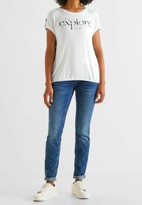 Bílý tričko s krátkým rukávem s textovým grafickým motivem „CESTUJ, ZKOUMEJ, ŽIJ“, kombinováno s modrými džínami a bílými teniskami. Ležérní střih a styl.
