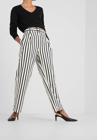 Un pull noir à col en V associé à un pantalon blanc court taille haute avec des rayures verticales noires et un détail de ceinture marron. Des escarpins noirs à bride arrière.