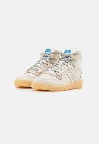 High-top sneakers in cream leather with perforated toe, gray laces, and a gum sole. Features blue logo tag. Subtle three stripes design.