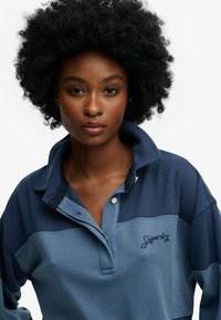 Młoda kobieta z naturalnymi włosami afro, ubrana w niebieską koszulkę rugby marki Superdry, patrząca przed siebie z neutralnym wyrazem twarzy na jednolitym tle.