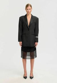 Ženska v prevelikem črnem blejzerju in črni prosojni midi krilu s črnimi špicastimi petami, stoji na belem ozadju.