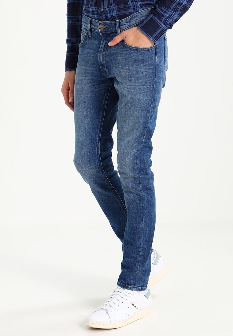 Slim-fit blue denim jeans with a faded wash, five pockets, and stitching details, worn with white sneakers on a plain background.