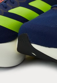 Kék sportcipők zöld csíkokkal, szövet felsőrésszel, lekerekített orral, textúrázott részletekkel és fekete gumitalppal, fehér középtalppal.