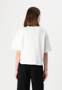 Tricou alb scurt, cu mâneci scurte, detalii cu cusături curbate și o croială lejeră, purtat cu pantaloni negri cu talie înaltă.