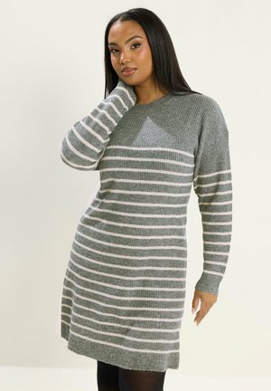 Kvinde med langt mørkt hår iført en grå og hvid stribet strik kjole med den ene hånd nær nakken, stående på en ensfarvet baggrund.