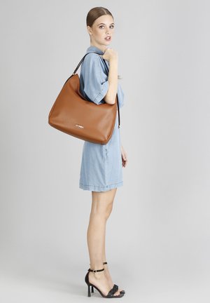 Bolso de mano de cuero marrón con una textura suave, de gran forma de hobo y correa para el hombro negra. Logo decorativo dorado en la parte frontal.