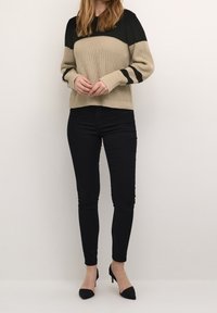 Pull en maille beige et noir avec un haut texturé, manches longues et deux rayures noires. Porté avec un jean skinny noir et des talons noirs.