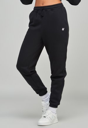 Persoon met zwarte joggerbroek met elastische boorden, witte sokken en witte sneakers, staand tegen een effen grijze achtergrond.