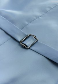 Ljusblått tyg med en mjuk yta, med en spänne i silverfärgad metall med öglor, säkrad av ett band i samma färg.