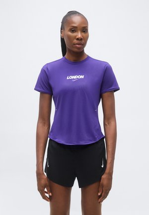 Γυναίκα που φοράει μωβ μπλουζάκι Nike με τη λέξη "London" και μαύρα σορτς Nike, στέκεται μπροστά σε απλό λευκό φόντο.