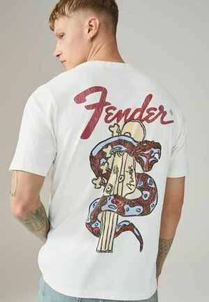 Junger Mann mit kurzem Haar trägt ein weißes T-Shirt mit rotem "Fender"-Schriftzug und einem Gitarrenkopf, um den sich eine rot-blaue Schlangengrafik auf dem Rücken windet.
